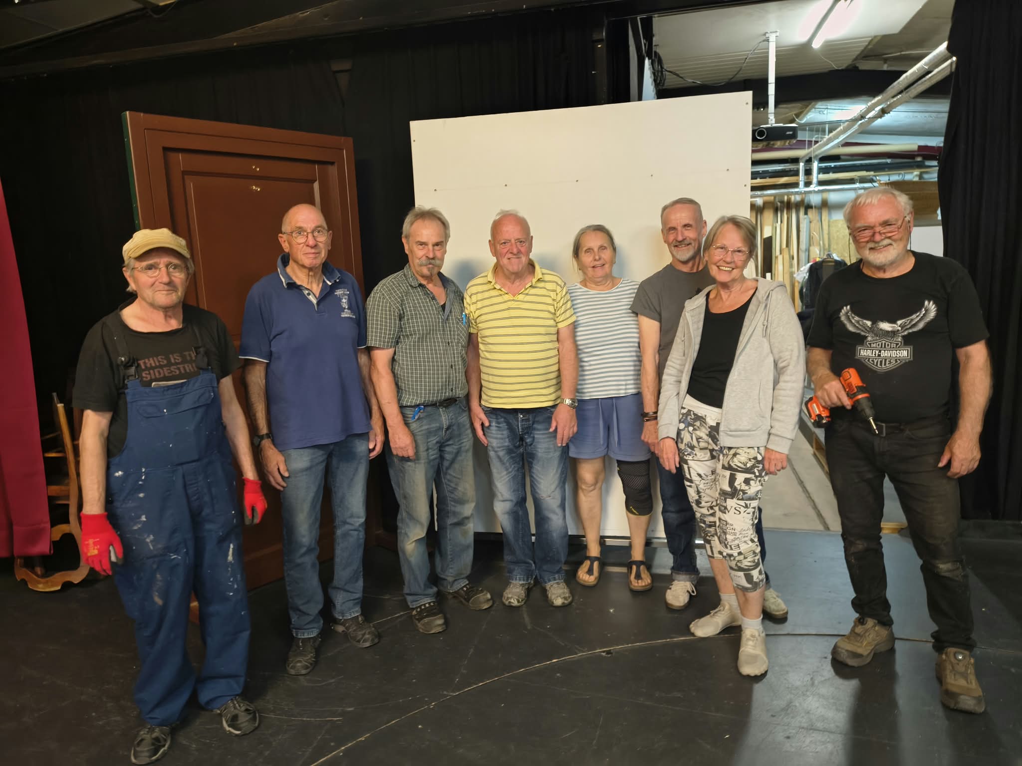 Acht Mitglieder des BAC-Theaters stehen mit Arbeitshandschuhen und Werkzeug vor den Bühnenbauteilen.